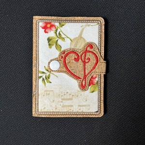 Puede incluir: Una cartera de corcho y tela con un diseño de corazón rojo y clave de sol. La cartera tiene un cierre de botón blanco.