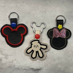 Op de afbeelding: Drie sleutelhangers met Mickey Mouse en Minnie Mouse figuren. De eerste sleutelhanger is zwart met rode stiksels en een blauwe knop. De tweede sleutelhanger is wit met zwarte stiksels en heeft Mickey Mouse' handschoen. De derde sleutelhanger is zwart met gele stiksels en heeft Minnie Mouse' hoofd met een roze strik.