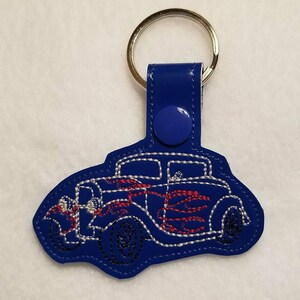 以下が含まれることがあります： 青いキーホルダー。刺繍されたクラシックカーのデザインで、赤い炎と白いステッチが施されています。キーホルダーには、シルバーの金属リングと青いスナップ留めが付いています。
