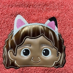 Könnte beinhalten: Gestickte braune Haare mit einem weißen Stirnband mit rosa Katzenohren auf einem korallenfarbenen Handtuch.