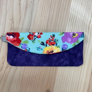 May include: A floral print clutch wallet with a teal top flap featuring red, purple, and yellow flowers. The bottom portion is a deep purple fabric. A small silver ring is attached to the side.