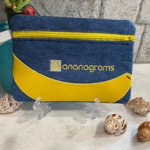 May include: A denim zippered pouch with a yellow banana design and the word "bananagrams" embroidered in yellow. The pouch has a yellow zipper and is displayed on a clear stand with seashells in the foreground.