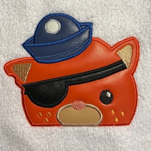 May include: Orange and brown cartoon animal face with a blue and black hat and an eye patch. The animal has a small nose and a big smile.