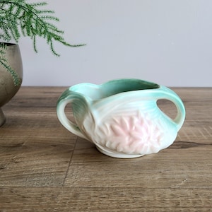 May include: A small, white ceramic planter with two handles. The planter is decorated with a pink flower design and a green glaze.