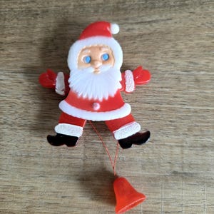 May include: A vintage Santa Claus Christmas ornament. The ornament is red and white with a white beard, blue eyes, and a red hat. A red bell hangs from a string attached to the ornament.