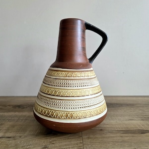 May include: A brown ceramic jug with a black handle. The jug has a cylindrical neck and a bulbous body decorated with horizontal bands of cream and gold geometric patterns. The base is brown, and the jug sits on a wooden surface.