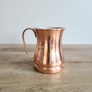 May include: A copper mug with a brass handle. The mug has a rounded body and a flared lip. It is sitting on a wooden surface.