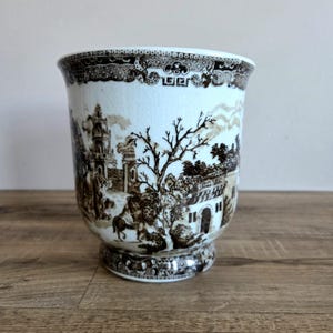 May include: A white and brown ceramic planter with a detailed landscape scene. The planter has a pedestal base and a decorative rim. The landscape features buildings, trees, and a horse and rider. The planter is on a wooden surface.