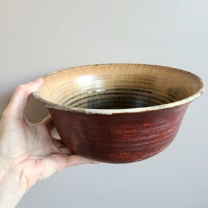 May include: A large, handmade ceramic bowl with a red exterior and a cream and brown interior. The bowl has a slightly flared rim and is glazed with a glossy finish.