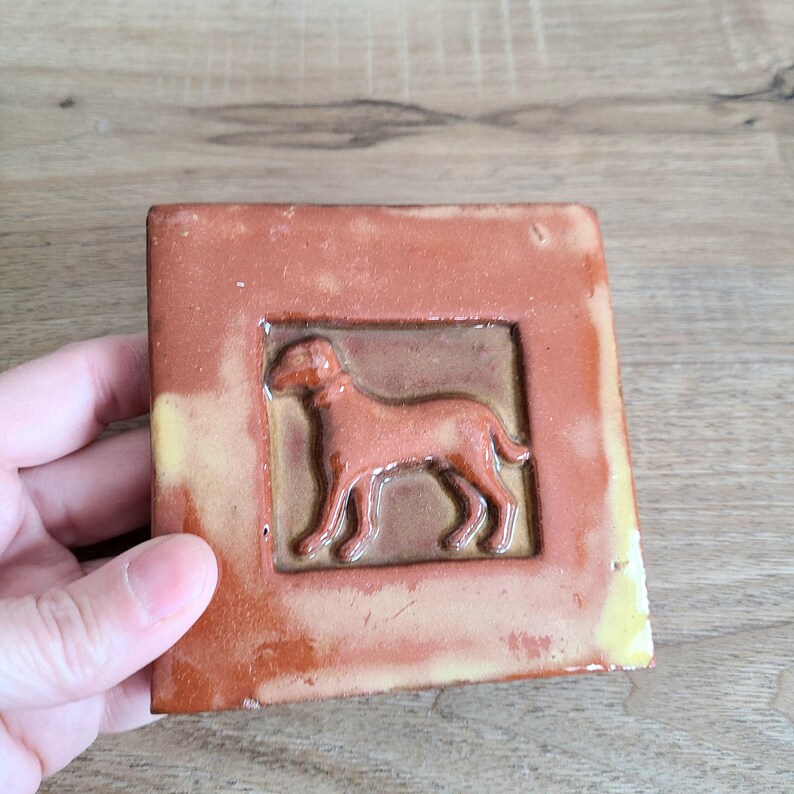 May include: A terracotta tile with a raised image of a dog in a brown and yellow frame.