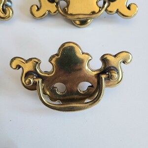 May include: An ornate cabinet pull in a gold tone with a decorative design and a curved handle.