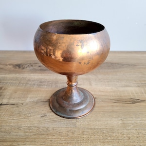 May include: A copper goblet with a round bowl and a flared base. The goblet has a rustic, aged appearance.