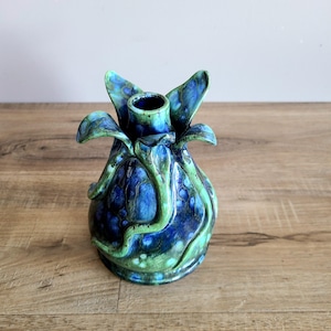 May include: A ceramic vase with an unusual, organic design. The vase has a bulbous body with a textured, mottled finish in shades of blue and green. The top has a flared, petal-like shape, and the overall aesthetic is reminiscent of a flower.