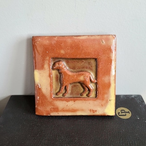 May include: A ceramic tile with a raised image of a brown dog in a standing position. The tile is a light brown color with a darker brown border.