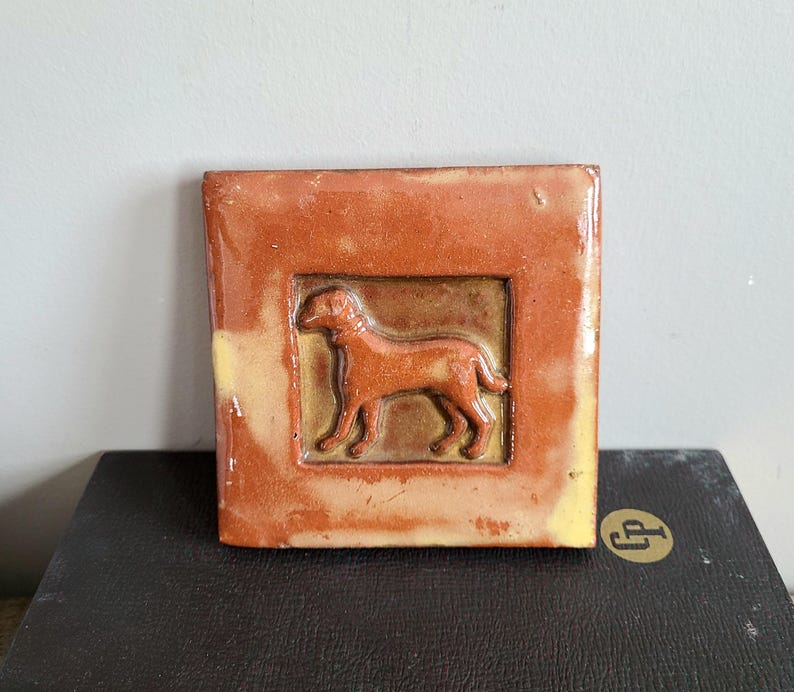 May include: A ceramic tile with a raised image of a brown dog in a standing position. The tile is a light brown color with a darker brown border.