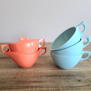 May include: A set of four pastel blue ceramic teacups stacked on top of each other. The cups have a rounded shape and a handle on the side. The cups are sitting on a wooden surface. Next to the cups is a set of two coral coloured ceramic teacups with a lid. The cups are also sitting on a wooden surface.