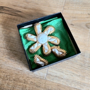 May include: A white flower-shaped brooch with gold accents and two matching earrings in a black box with green velvet lining.