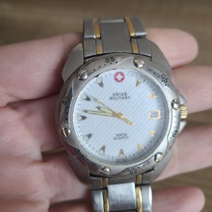 Peut inclure: Une montre Swiss Military bicolore argent et or avec un cadran blanc, des aiguilles noires et une fenêtre de date. La montre est étanche à 10 ATM et possède un mouvement à quartz.