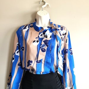 May include: A blue and beige patterned blouse with a large bow tie. The blouse has a long sleeve and a relaxed fit.