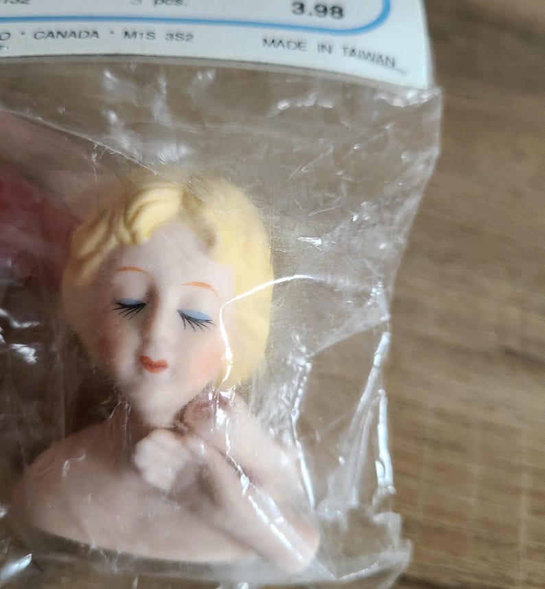 May include: A porcelain doll head with closed eyes and blonde hair. The head is in a plastic bag with a price tag that reads "3.98 Made in Taiwan".