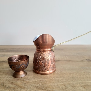 May include: A copper coffee pot and small cup on a wooden surface. The pot has a long handle and floral pattern. The cup has a decorative border. Both items have a shiny, metallic finish.