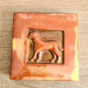 May include: A brown ceramic tile with a raised image of a dog in a brown frame. The tile has a rustic, handmade look.