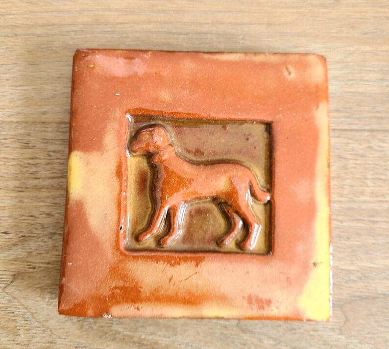 May include: A brown ceramic tile with a raised image of a dog in a brown frame. The tile has a rustic, handmade look.