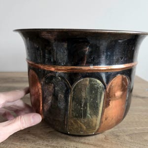 May include: A vintage metal planter with a dark, aged finish. The planter has a copper band and decorative panels in copper, brass, and bronze. The rim flares outwards, and the shape is rounded.
