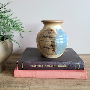 May include: A ceramic vase with a blue and brown glaze. The vase is sitting on top of two books. The top book is brown and has the title "Traveling Toward Sunrise" on the spine. The bottom book is pink and has the title "Fairy Tales" on the spine.