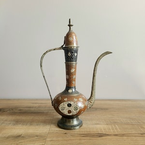 May include: A decorative brass teapot with an ornate design. The teapot has a round body with a long, curved spout and a handle. The teapot is decorated with intricate floral patterns in shades of gold, orange, and black.