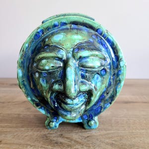 Peut inclure: Une sculpture en céramique représentant un visage souriant aux yeux fermés. Le visage est rond avec un glaçage bleu et vert. La sculpture a une forme circulaire avec un bord surélevé et trois petits pieds.