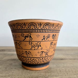 May include: A terracotta-coloured ceramic bowl featuring black painted details. The exterior displays horizontal bands of stylised animal figures, including lions and other creatures, interspersed with decorative floral and geometric patterns.