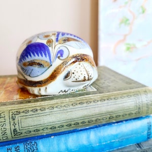 May include: A ceramic ornament with a rounded form, decorated with blue, white, and brown floral patterns. The ornament is placed on a stack of old books. The word "Mexico" is inscribed on the ornament.