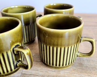 May include: Four olive green ceramic mugs with vertical ribbed detailing. Each mug has a rounded handle and a glossy finish. The mugs are arranged on a light brown wooden surface.