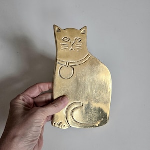 Peut inclure: Un objet décoratif en laiton en forme de chat. Le chat est assis, portant un collier avec une étiquette circulaire. Le visage du chat comporte des détails gravés pour les yeux, le nez et les moustaches. L'objet est de couleur dorée.