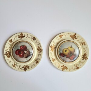 May include: Two round, cream-coloured plates with gold painted floral designs. Each plate has a smaller, round, glass-covered picture of fruit. One plate features red tomatoes and walnuts, the other features a yellow apple and strawberries.