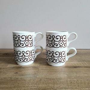 May include: Four white ceramic mugs with brown floral designs. The mugs are stacked in pairs, with the top mugs having handles.