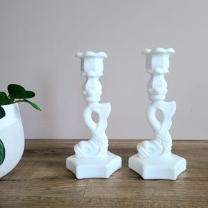 May include: A pair of white dolphin-shaped candle holders. Each candle holder features a square base, a detailed dolphin design, and a scalloped top. The candle holders are made of a smooth, opaque material and are on a wooden surface.