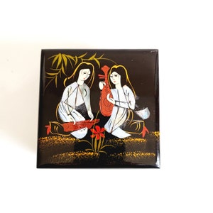 May include: A black wooden box featuring a painted scene of two women playing traditional Vietnamese instruments. The women are dressed in white and are surrounded by gold and red flowers.