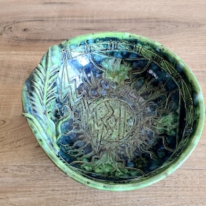 May include: A handcrafted ceramic bowl with a distinctive design. The bowl has a textured surface with a green and blue glaze. The interior features an intricate pattern with a sun-like design and etched symbols. The rim has a leaf-like detail.
