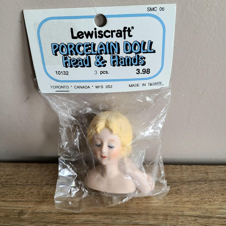 May include: A set of three porcelain doll heads and hands. The heads are white with painted features and blonde hair. The hands are white and have a slight pink tint. The packaging is blue and white with the text "Lewiscraft Porcelain Doll Head & Hands 10132 3 pcs. 3.98 Made in Taiwan".