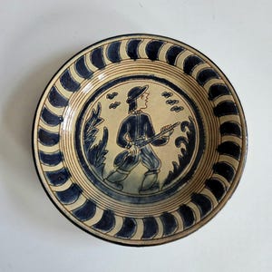 May include: A round ceramic plate in blue and cream. The plate's central design depicts a person in blue clothing with a rifle, set amidst foliage and birds. The rim is decorated with a repeating blue and cream pattern.
