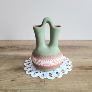 May include: A green and brown ceramic vase with two openings and a handle. The vase features a traditional Native American design.