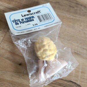 May include: A set of three porcelain doll head and hands for crafting. The head is yellow and the hands are a light skin tone. The packaging says "Lewiscraft T&Ecirc;TE et MAINS de Porcelaine 10132 3 pcs. 3.98 TORONTO CANADA M1S 3S2 FABRICUS EN TAIWAN"
