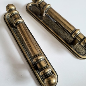 May include: Two antique bronze door handles. Each handle has a long, rectangular backplate with a textured surface and a central, cylindrical grip. The grips are decorated with rings and topped with a rounded finial. The handles are made of metal.
