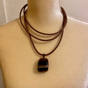 Puede incluir: Un collar de cordón de cuero marrón con un diseño en capas y un colgante hecho de una piedra rayada de marrón oscuro, negro y blanco.
