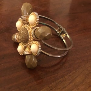 May include: A gold-tone bangle bracelet with a large, ornate, green and white centerpiece. The centerpiece features a floral design and is surrounded by smaller green and white beads.