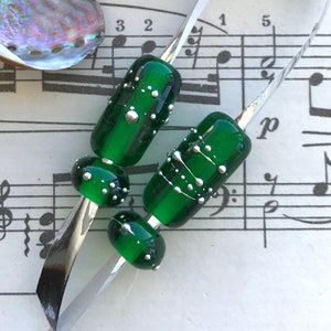 May include: Four green glass beads with silver accents. The beads are on a silver metal surface. The beads are round and have a speckled design.