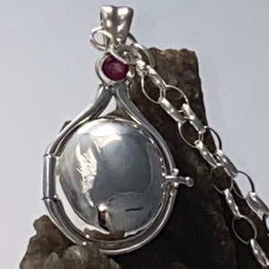 Puede incluir: Collar con dije de plata con una piedra preciosa roja y una cadena. El dije está abierto y tiene una superficie lisa y reflectante.