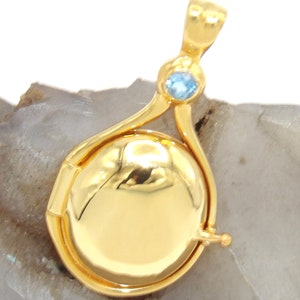 May include: A gold locket pendant with a blue gemstone accent. The locket is open and has a round, shiny interior.